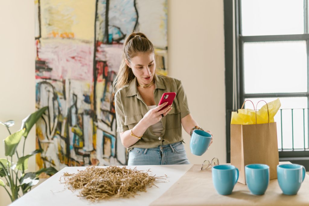 O femeie folosește telefonul în timp ce stă în fața unei mese cu cești de cafea și alte obiecte. (Bani din dropshipping)